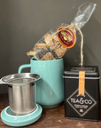 Tea-making set with teal mug, tea leaves, stainless steel infuser, and black tea canister on a wooden surface.