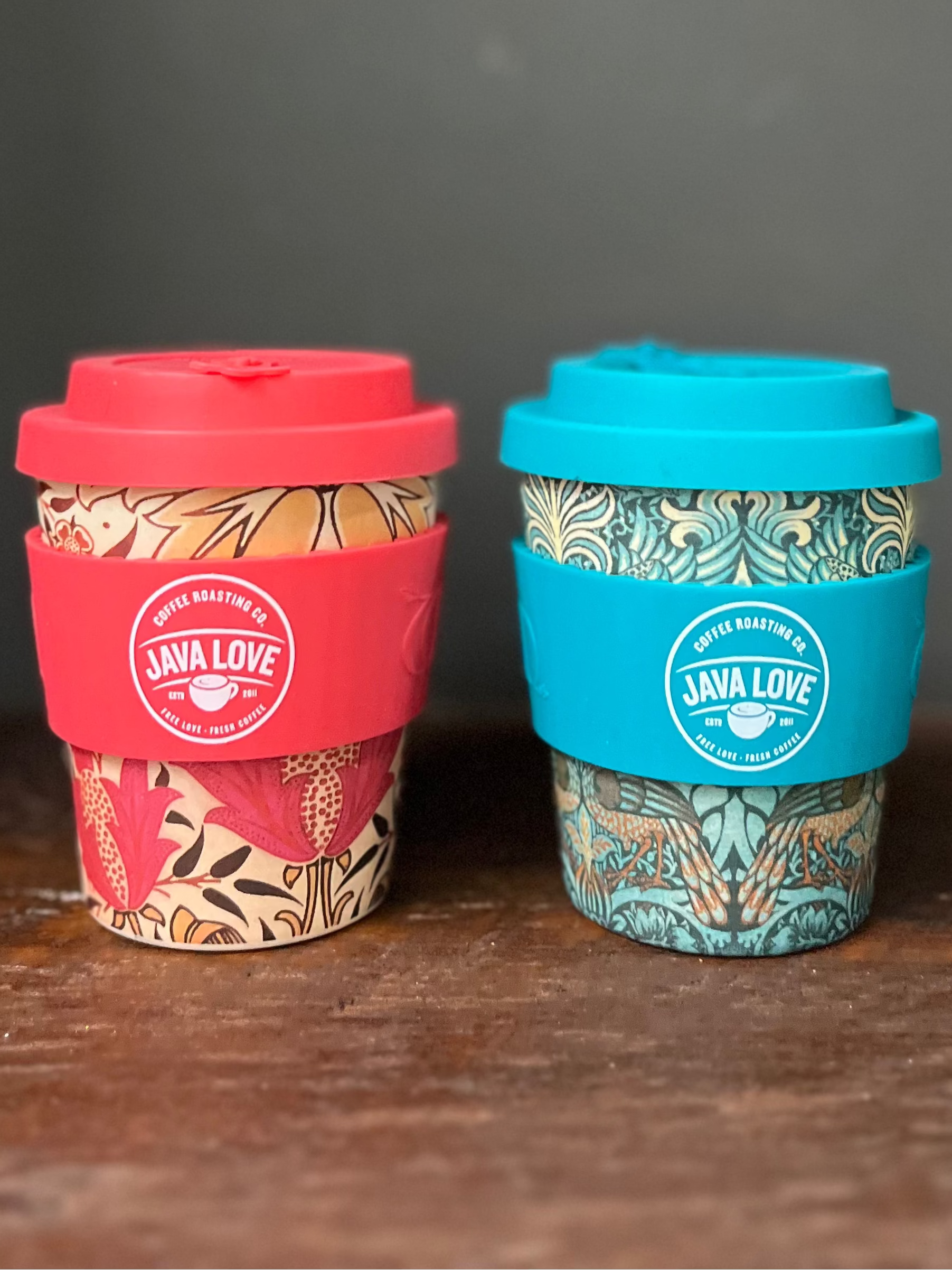 Two reusable coffee cups with colorful patterns and red and blue lids on a wooden surface.