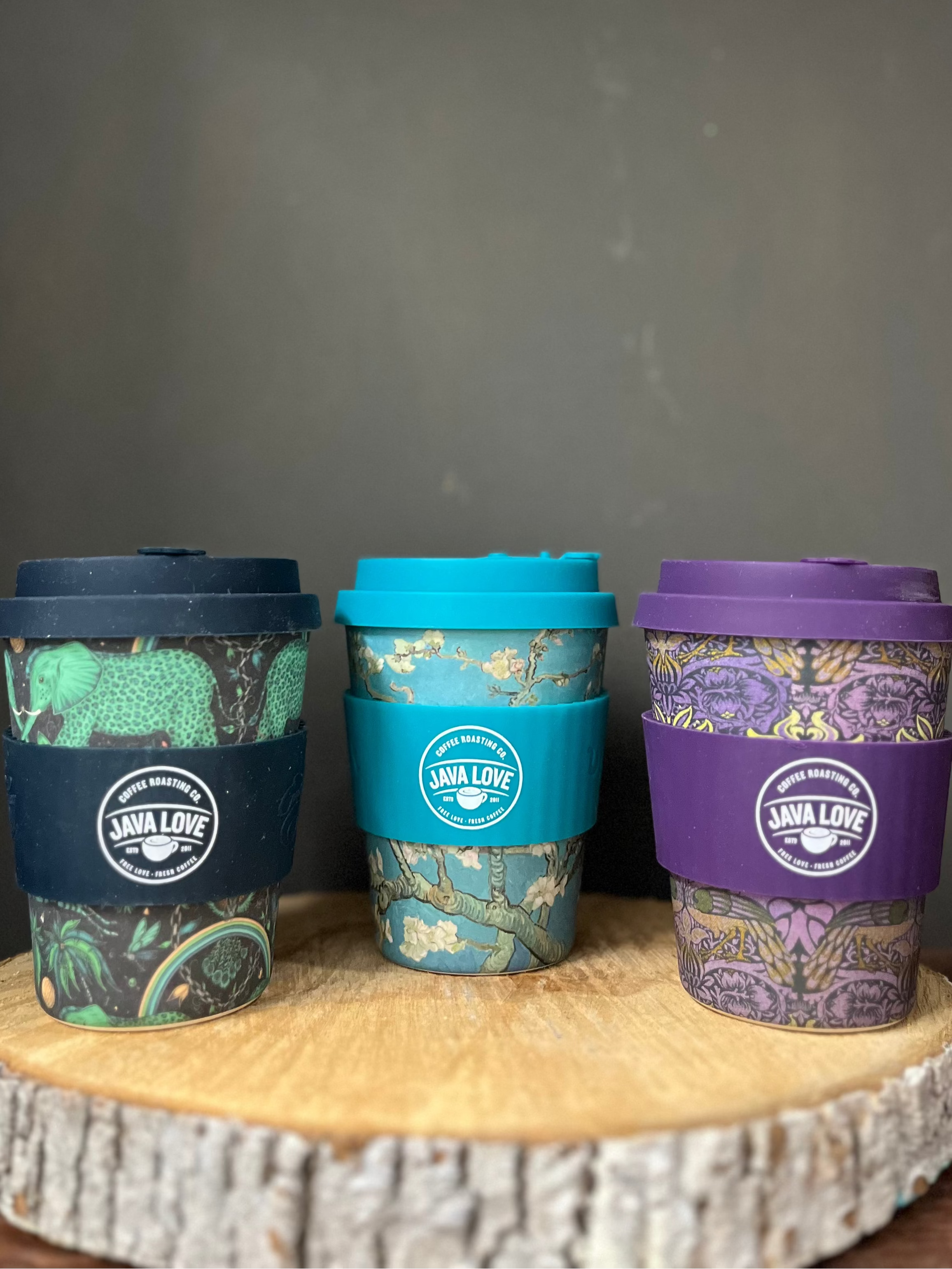 Three reusable coffee cups with different color lids on a wooden stand against a dark background